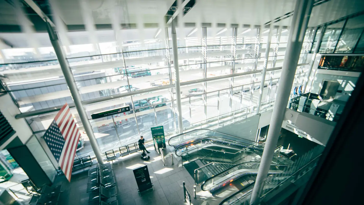 A bright, spacious airport terminal featuring expansive glass walls, escalators, and American flag.