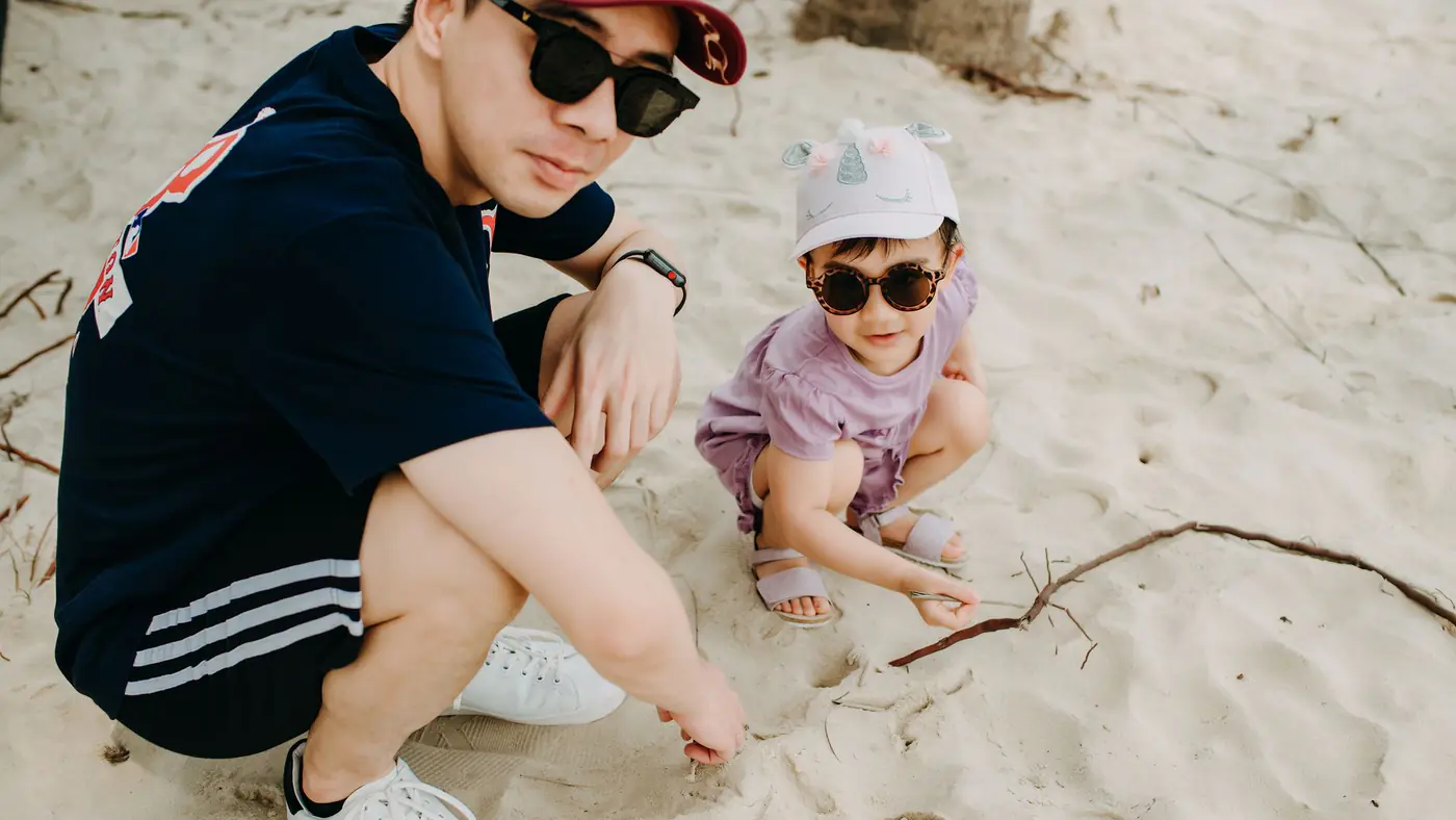 A father and his child playing on a sandy beach, both wearing sunglasses and casual summer attire.