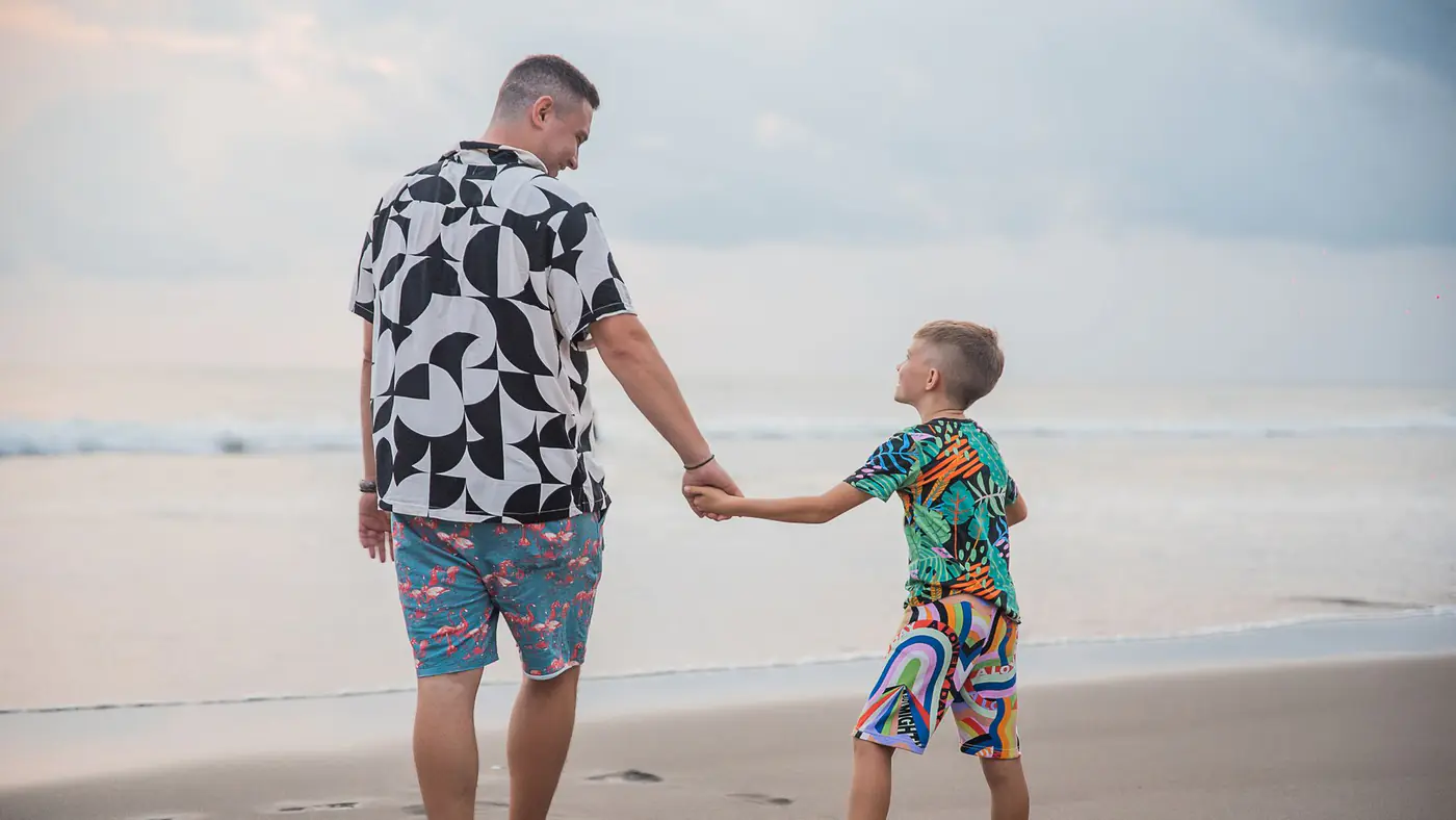 A father and son holding hands, walking on a sandy beach during a serene sunset.