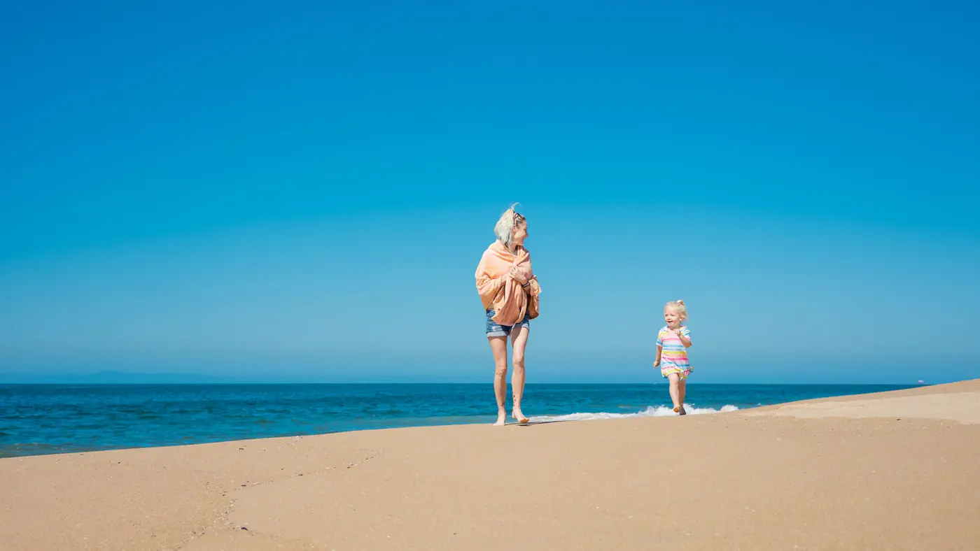 A mother and daughter walking on a sunny beach with a clear blue sky.
