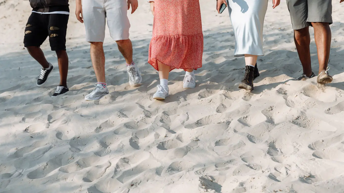 A diverse group of people walking on a sandy beach, showcasing summer fashion and casual footwear.