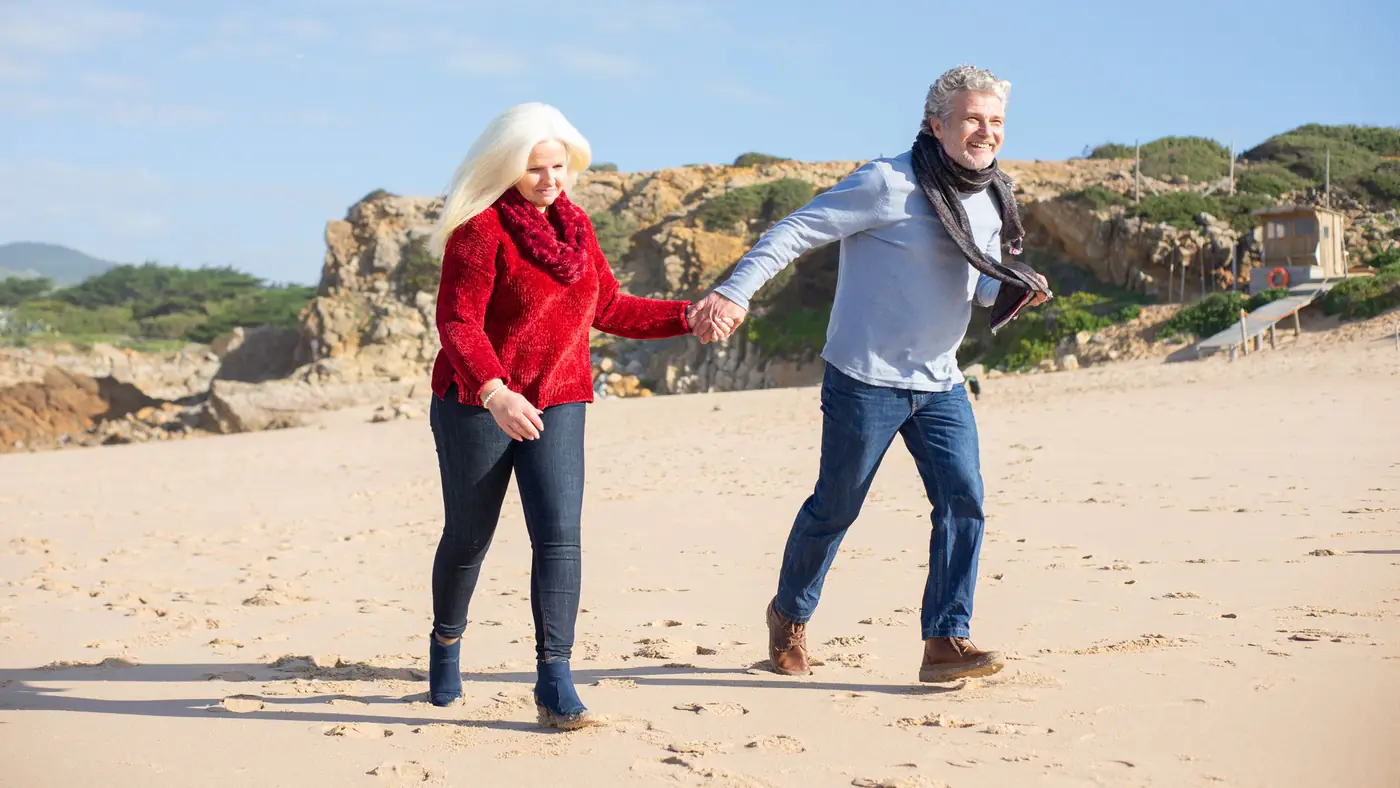 An elderly couple holding hands and walking on a sunny Portuguese beach, showcasing love and companionship.